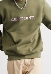 Oliivinvihreä pusero pyöreällä pääntiellä, jossa on vaaleanpunainen kirjailtu "carhartt"-logo. Sileä pinta, resorit hihansuissa ja helmassa.