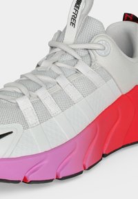 Άσπρο αθλητικό παπούτσι από δίχτυ με λευκά κορδόνια, ετικέτα "NIKEFREE" και χοντρή σόλα με διαβάθμιση χρώματος από ροζ σε κόκκινο.