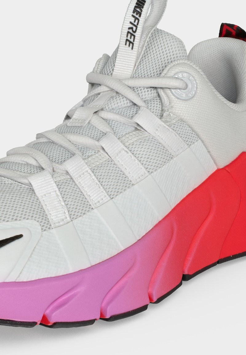 Zapatilla deportiva blanca de malla con cordones blancos, lengüeta "NIKEFREE" y una suela gruesa con degradado que cambia de rosa a rojo.