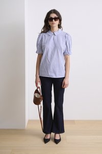 Blusa a righe a maniche corte con colletto, abbinata a jeans svasati scuri. Gli accessori includono occhiali da sole oversize e una piccola borsa marrone.