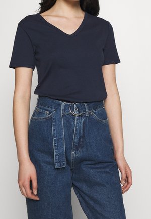 Frau trägt ein marineblaues V-Ausschnitt kurzärmeliges Oberteil, das in hoch taillierte blaue Jeans aus Denim mit Stoffgürtel und silberner Schnalle gesteckt ist.