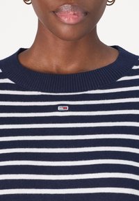 Sweter w biało-niebieskie paski z żebrowanym okrągłym dekoltem i małą łatą z logo na piersi. Materiał wydaje się miękki i dziany.