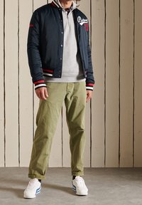 Giacca da bombardiere navy con accenti rossi e bianchi, felpa grigia con cerniera, pantaloni verde oliva e scarpe bianche con strisce blu.