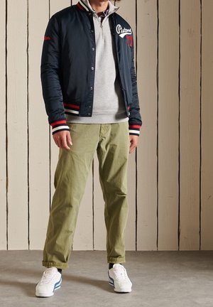 Navy bomber jacket with red and white accents, grey zip-up sweatshirt, olive green pants, and white sneakers with blue stripes.