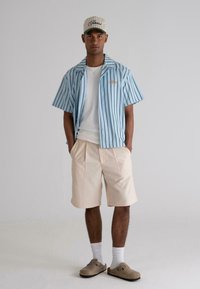 Short-sleeve blue striped shirt with a gold logo, white t-shirt underneath, light peach shorts, white socks, and brown slip-on shoes.