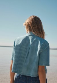 Chemise en denim à manches courtes bleu clair avec des rayures verticales, dotée d'un empiècement au dos, d'un col et d'un design court. Texture lisse.