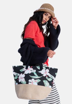 Frau mit Strohhut, rotem Hemd und schwarzem Pullover trägt eine große Blumentasche und posiert vor einem weißen Hintergrund.