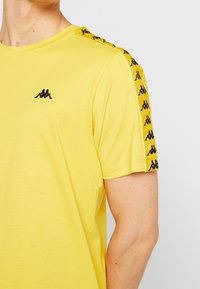 Muž oblečený v žluté košili s krátkým rukávem, se černými logo pásky na ramenou a malým černým logem na hrudi.