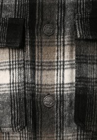 Țesătură flanelată în carouri gri și bej, având două butoane metalice și două buzunare cu detalii negre și un finisaj moale, texturat.
