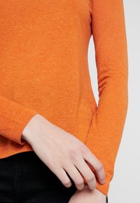 Eine Person trägt ein oranges Langarmshirt und passt mit einer Hand den Ärmel nahe dem Handgelenk an, vor einem schlichten Hintergrund.