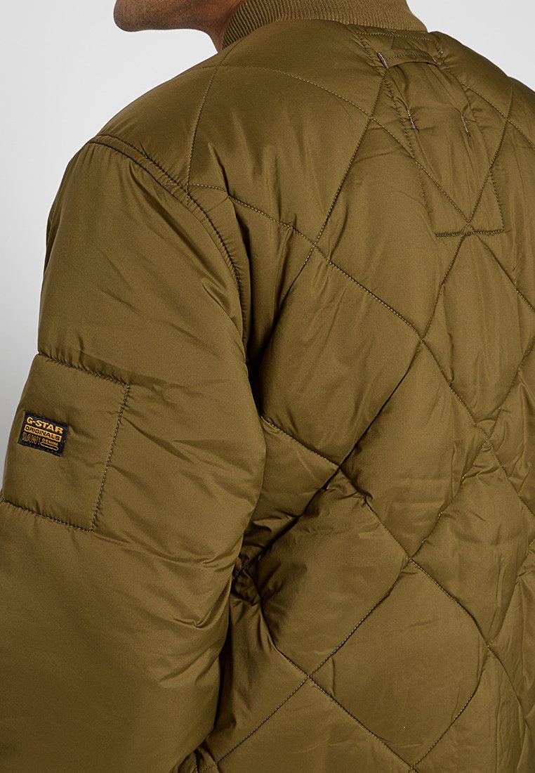 Mann trägt eine olivgrüne Steppjacke mit geripptem Kragen und einem G-Star Originals-Logo-Patch am Oberarm.