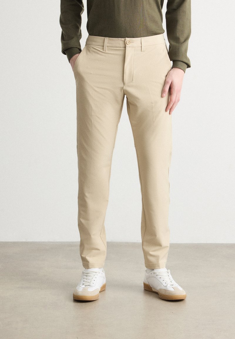 Beige trousers made of soft fabric, featuring a slim fit, front pockets, and a button closure, paired with white sneakers and a green top.