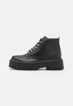Schwarzer Leder-Stiefelette mit dicker Profilsohle und schwarz-weißen, markenbezogenen Schnürsenkeln vor weißem Hintergrund, Seitenansicht.