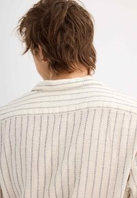 Camisa beige texturizada con rayas verticales azules, fabricada en un suave tejido entrelazado, y con un collar en la parte posterior, que muestra un escote abierto.
