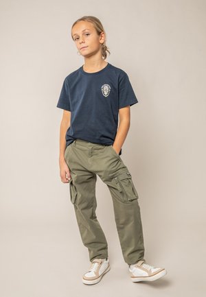 Jeune personne debout, les mains dans les poches d'un pantalon cargo olive, portant un t-shirt graphique bleu marine et des baskets blanches beige, sur un fond uni.