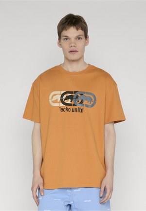 Fiatal férfi rövid hajjal, narancssárga Ecko Unltd pólót visel, amelyen három hippo logó látható, világoskék rövidnadrággal és fehér feliratokkal.