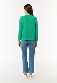 Grüner gestrickter Pullover mit langen Ärmeln, strukturiertem Webmuster und Rundhalsausschnitt; kombiniert mit hellblauen Schlagjeans und silbernen Sneakern.