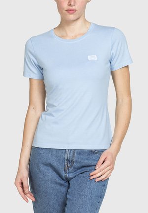 Donna che indossa una T-shirt azzurra a maniche corte con un piccolo logo bianco sul petto, abbinata a jeans di denim blu.