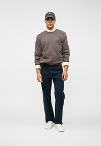 Uomo che indossa una felpa grigia sopra una maglietta gialla, pantaloni blu scuri, scarpe da ginnastica bianche e un berretto scuro, in piedi contro uno sfondo chiaro e uniforme.