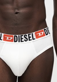 Gros plan d'une personne portant un slip blanc avec une large ceinture affichant le logo de la marque Diesel en noir et orange.