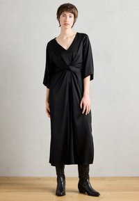 Musta, polvipitkä mekko syvällä V-aukolla, solmukohtaisella vyötäröllä ja löysillä hihoilla. Mallilla on jalassaan mustat korkokengät. Tasainen kangas.