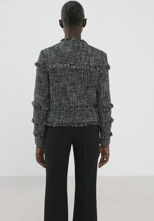 Persona con el cabello recogido en un moño, vestida con una chaqueta con flecos texturizados en blanco y negro y pantalones negros, de pie de espaldas contra un fondo liso.