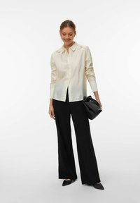 Chemise boutonnée beige clair à manches longues, avec des détails brodés sur le col ; associée à un pantalon noir à jambes larges et un sac noir.
