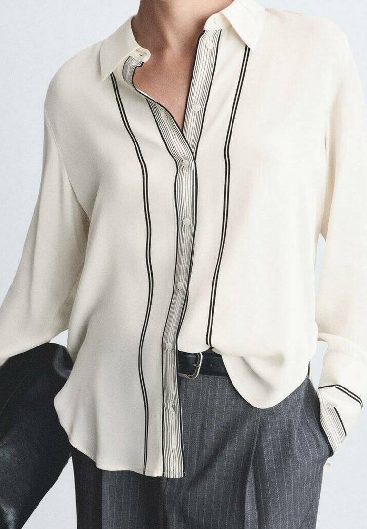 Blouse crème à manches longues avec des rayures verticales noires et des détails de boutons, rentrée dans un pantalon gris à fines rayures avec une ceinture noire.