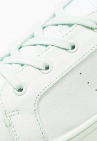 Białe zamszowe sneakersy o teksturowanej powierzchni, z jasnymi miętowymi sznurówkami i okrągłymi oczkami. Posiadają minimalne perforacje zapewniające wentylację.
