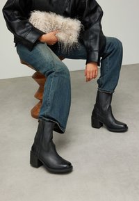 Bottines en cuir noir avec bout carré et talon épais, associées à un jean bleu en denim et une pochette texturée de couleur claire.