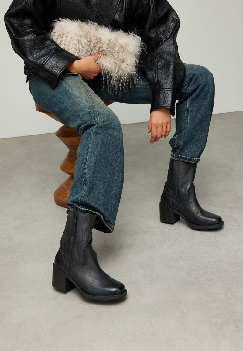 Bottines en cuir noir avec bout carré et talon épais, associées à un jean bleu en denim et une pochette texturée de couleur claire.