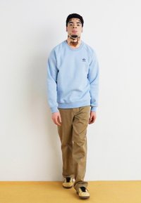 Svetlo modra majica s kapuco z rebrastimi manšetami, v kombinaciji z bež hlačami in črnimi supergami z rumenimi poudarki. Preprost dizajn, srednji kroj.