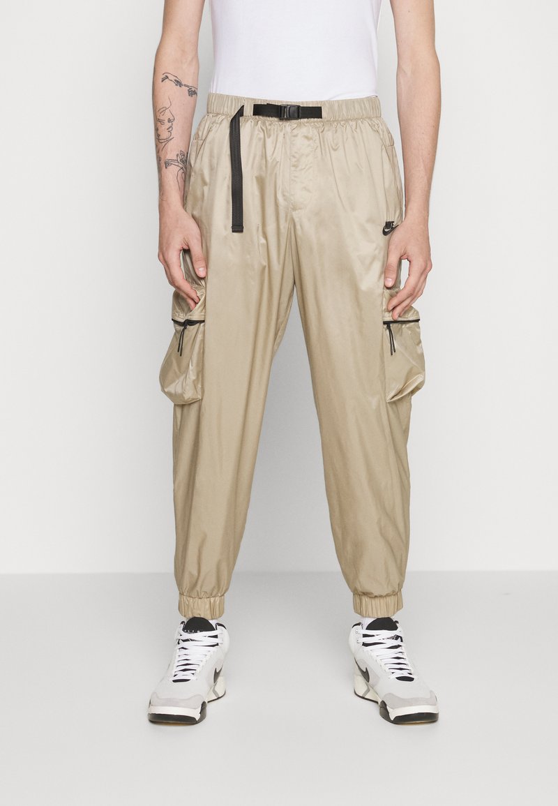 Pantalons cargo beiges, légers, avec une taille élastique, des chevilles resserrées et des poches latérales zippées. Accompagnés de baskets blanches et noires.