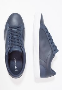 Baskets en cuir bleu marine avec semelles en caoutchouc blanc. Caractéristiques : texture lisse, bout rond et six œillets pour les lacets. Logo de la marque visible.