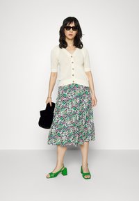 Cardigã de malha creme com mangas curtas e botões dourados, combinado com uma saia midi floral verde e sandálias de salto bloco verde. Acessório: mala preta.