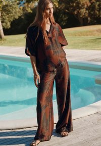Haut et pantalon à motif floral marron en tissu léger, avec des jambes larges et une coupe décontractée. Le mannequin se tient près d'une piscine.