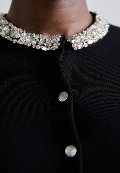 Svart cardigan med en smyckad krage i klara och silverfärgade accenter, texturerad stickad tyg och silverknappar framtill.