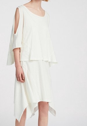 Žena nosící volné bílé šaty s výřezy na ramenou a asymetrickým lemem, stojící na jednobarevném bílém pozadí.