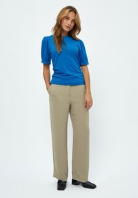 Mėlynas trumpų rankovių viršus su prigludusia forma, derinamas su smėlio spalvos plačiais kelnėmis ir juodais kvadratiniais bateliais ant neutralios fono.