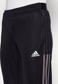 Černé sportovní kalhoty s texturovaným povrchem, s bílým logem Adidas a třemi vertikálními šedými pruhy po stranách. Součástí je elastický pas.
