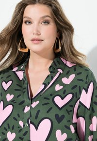 Groene blouse met een hartpatroon in roze en zwart, met een puntkraag en knoopsluiting. Model draagt grote gouden oorbellen.