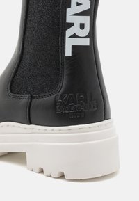 Bottine en cuir noir avec panneau en tissu élastique, impression de logo blanche, marquage en relief et semelle blanche épaisse avec un motif texturé.