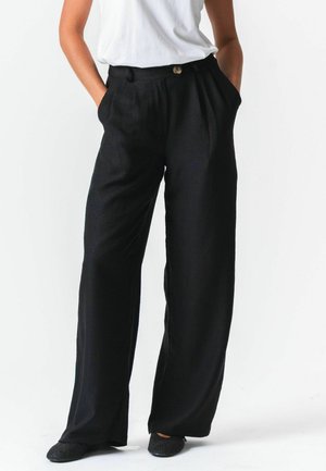 Person trägt locker sitzende, schwarze, hoch taillierte Hose mit Falten, ein weißes, in die Hose gestecktes T-Shirt, Hände in den Taschen und schwarze flache Schuhe.