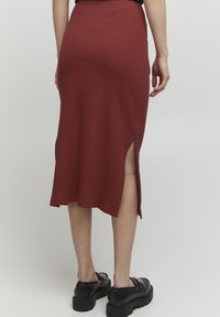 Rippierte Midirock in Dunkelrot mit seitlichen Schlitzen. Hat ein körperbetontes Design und reicht bis unter das Knie. Kombiniert mit schwarzen Plateauschuhen.
