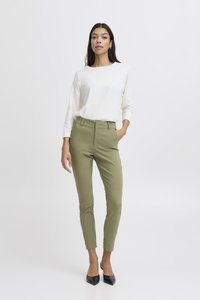 Femme debout avec la main dans la poche, portant un haut blanc à manches longues, un pantalon slim vert olive et des escarpins noirs à bouts pointus.