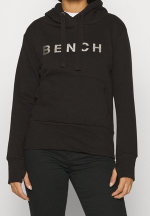 Osoba ubrana w czarną bluzę z kapturem z napisem "BENCH" w srebrnych literach, w połączeniu z czarnymi spodniami, ręce swobodnie opuszczone wzdłuż ciała.