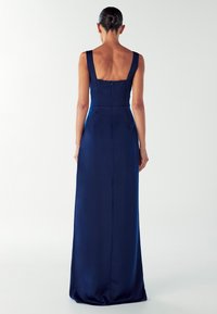 Moteris su suku į buną, vilkinti be rankovių, grindis siekiančią tamsiai mėlyną suknelę, stovi nugara pasukusi priešais paprastą baltą foną.