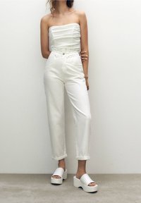 Weißes, trägerloses Oberteil mit gerafftem Design, kombiniert mit hoch taillierten weißen Jeans aus Denim und weißen Plateausandalen. Schlichte, klare Ästhetik.