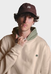Sudadera beige con capucha interior verde, con un logo de elefante verde bordado. Gorra marrón oscuro con un logo de elefante a juego en la parte delantera.