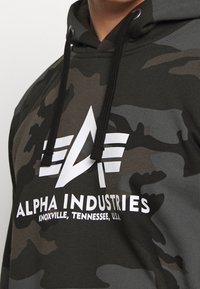 Kamouflage-hoodie i mörka gröna och grå nyanser, med en stor vit logotyp "ALPHA INDUSTRIES" och dragsko. Materialet verkar mjukt och texturerat.